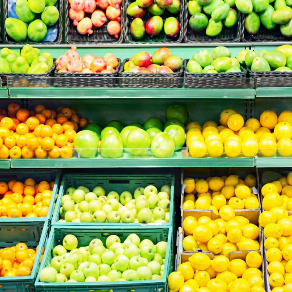 Rows of fresh produce including oranges, lemons, limes, apples, mangoes, and potatoes on market shelves.