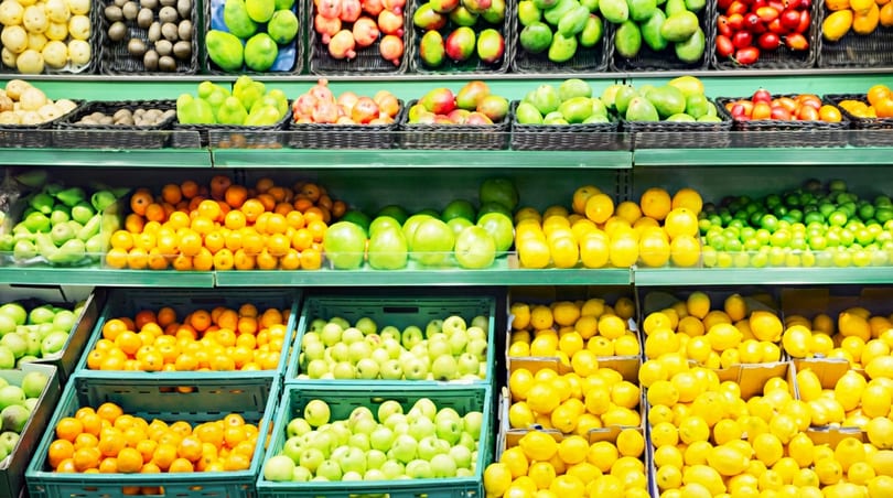 Rows of fresh produce including oranges, lemons, limes, apples, mangoes, and potatoes on market shelves.