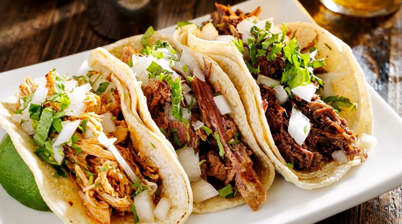 Three corn tortilla tacos filled with shredded chicken and beef, topped with fresh onions and cilantro.