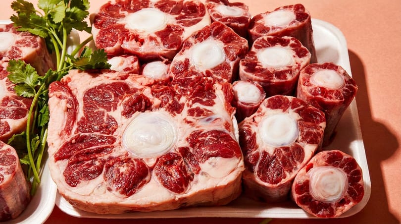 Raw oxtail pieces and beef shanks with fresh parsley on white trays.