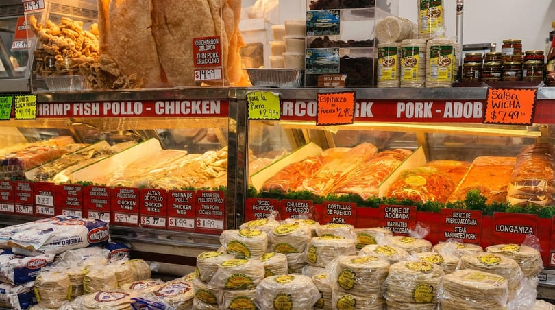 Latin American deli counter with prepared meats, large pork cracklings, and stacks of fresh tortillas.